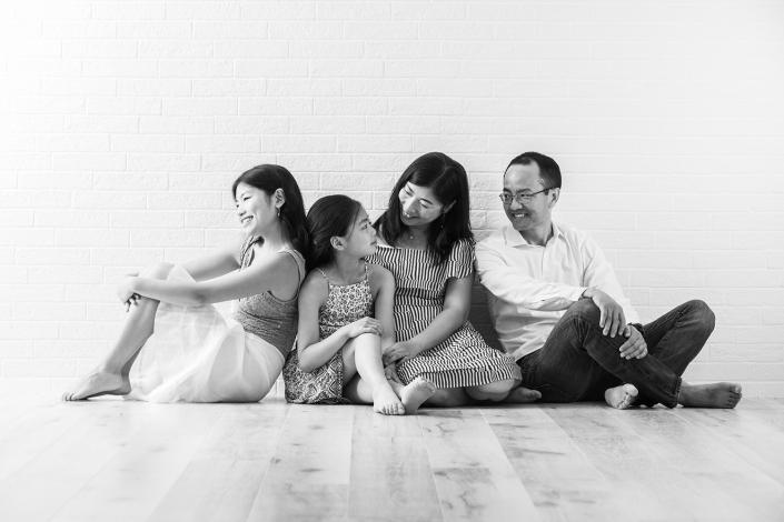 Timeless black and white portrait of an Asian family gathered indoors, sitting on the floor against a brick wall backdrop, radiating warmth and togetherness.