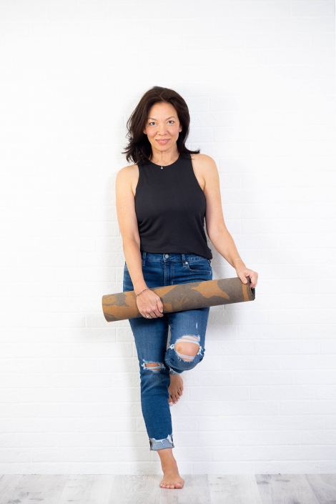 Confident female yoga teacher with a mat, dressed in jeans, posing against a white brick wall with a sense of calm and expertise.