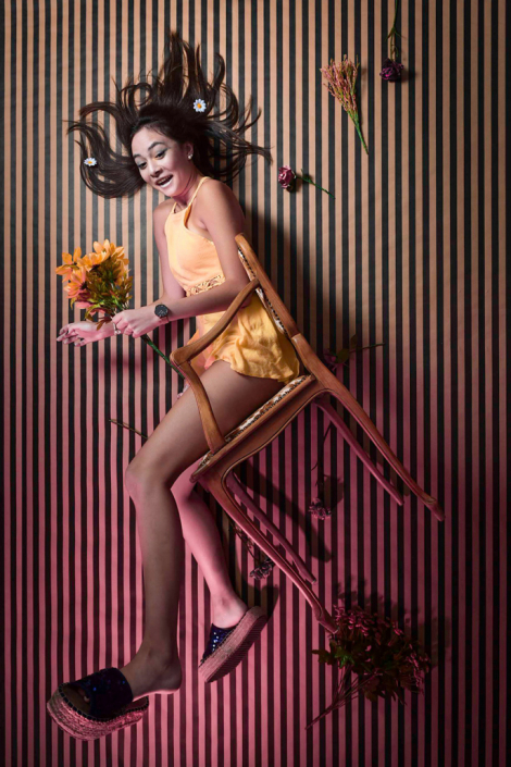 Artistic in-studio shot: girl seated on a chair, creatively mimicking a falling gesture amidst a vibrant floral environment.
