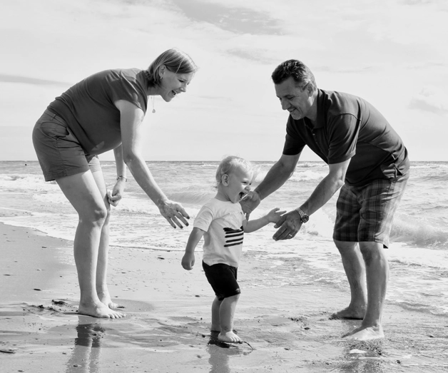 Delighted mother and father joyfully assisting their son as he takes his first steps on the picturesque beach of Prince Edward Island.